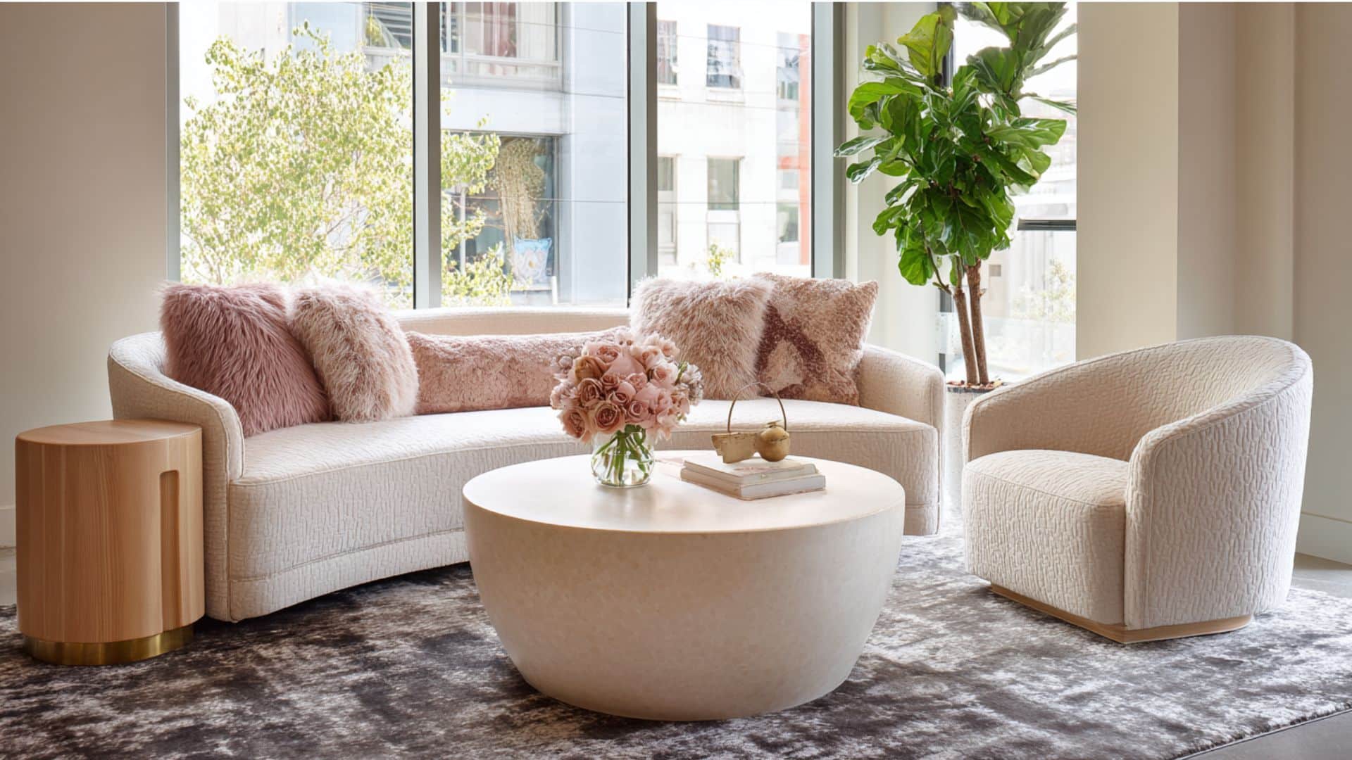 Modern living room with a white sofa, pink pillows, round coffee table, and a large plant by the window.