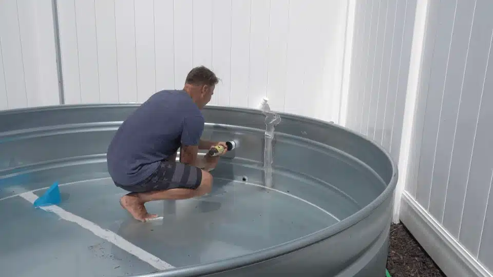 man inside a galvanized stock tank using a power drill and hole saw to create a hole in the side wall (1)