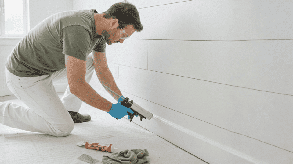 A person wearing safety goggles and gloves applying caulk along the trim of a shiplap half wall for a clean finish