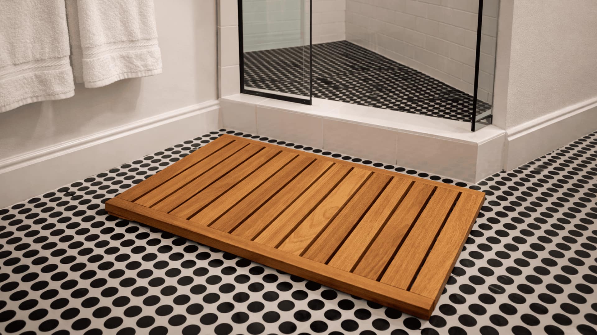teak wood shower mat on black and white penny tile floor beside glass shower and white walls