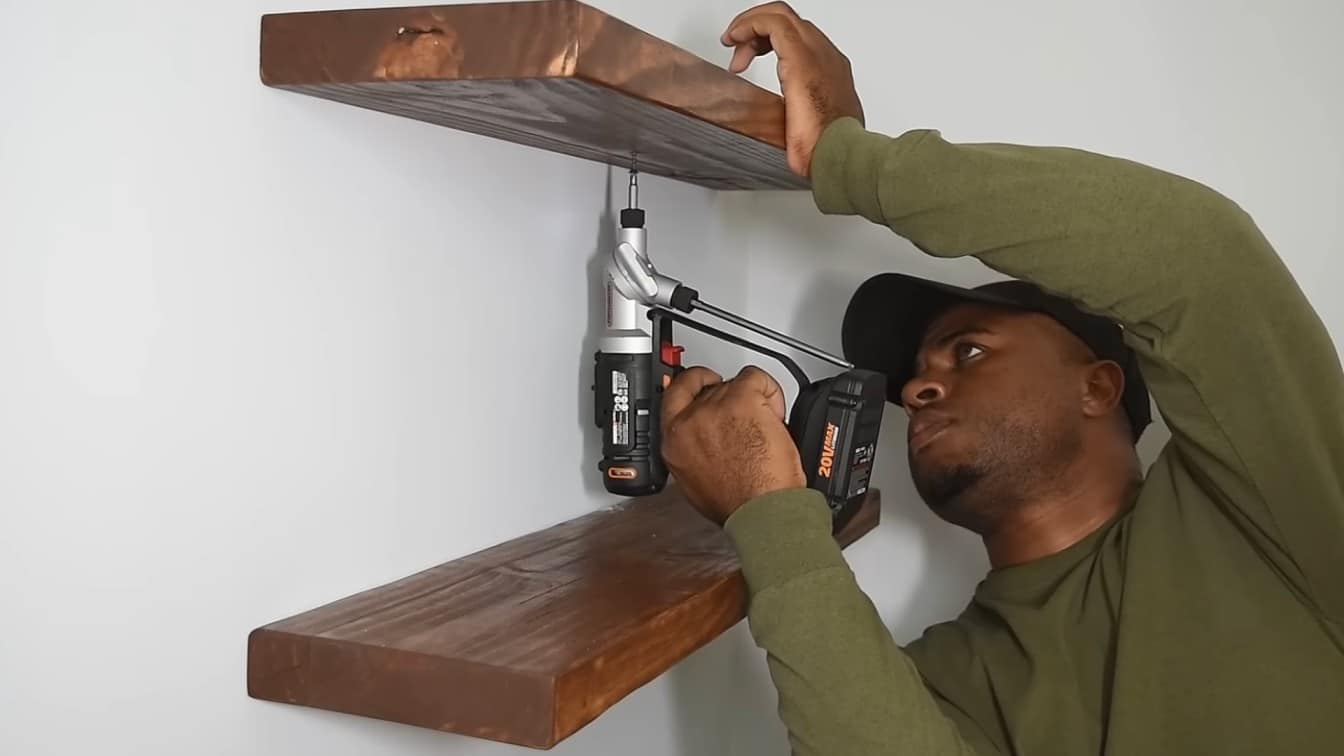 a man in a green shirt uses a power drill to secure two wooden shelves to a wall