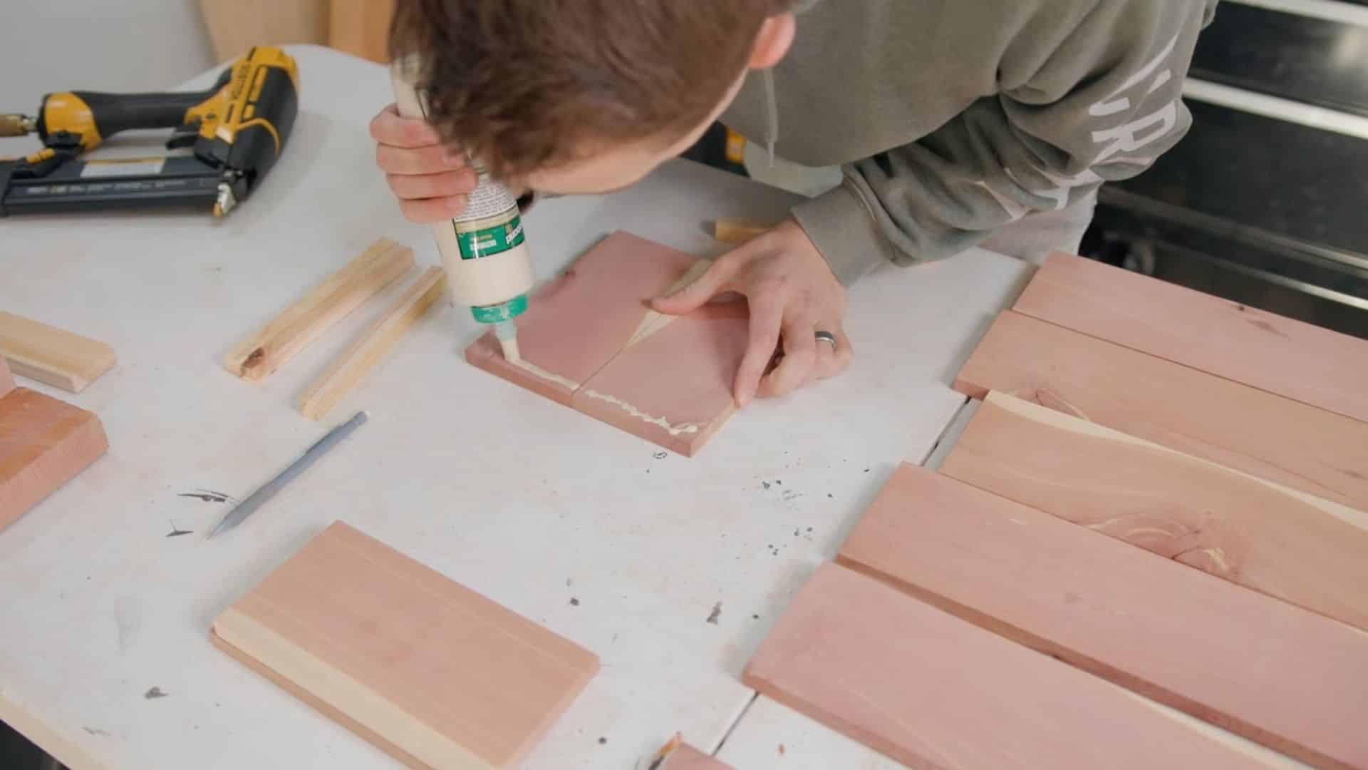 a person applying wood glue to the edge of a wooden board as part of assembling a flower box, with several other wooden pieces and tools visible on the work surface