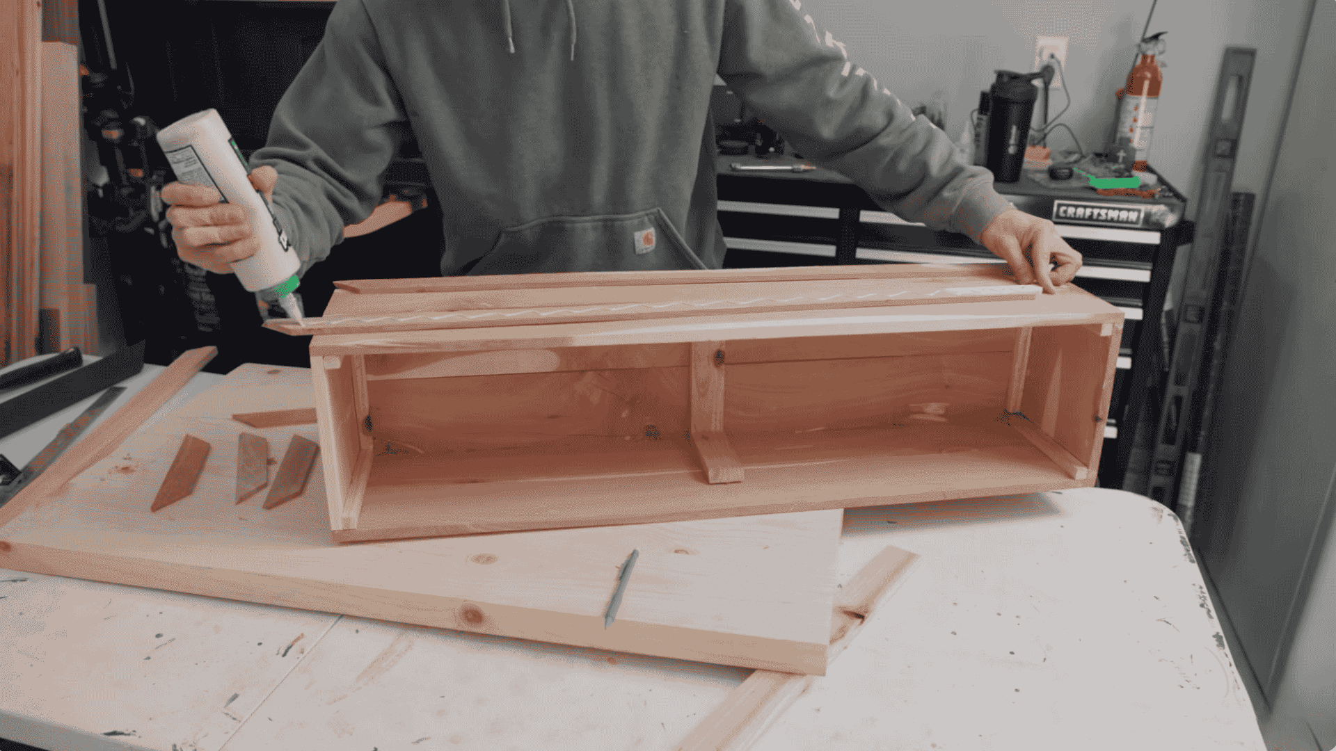 a person applying wood glue to the edge of a wooden piece while assembling a flower box, with the box partially assembled and other wood pieces laid out on the work surface