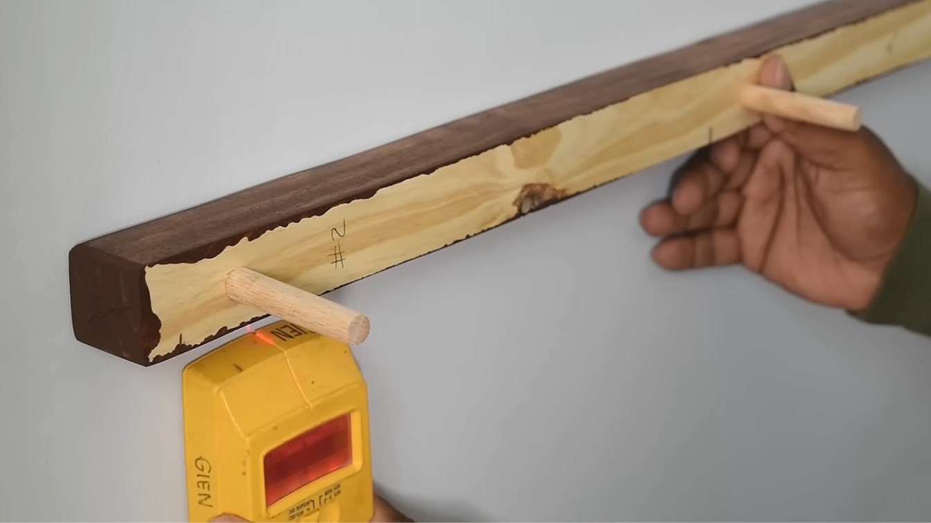 a person holds a wooden rail with dowels against a wall, using a yellow stud finder to locate the wall studs