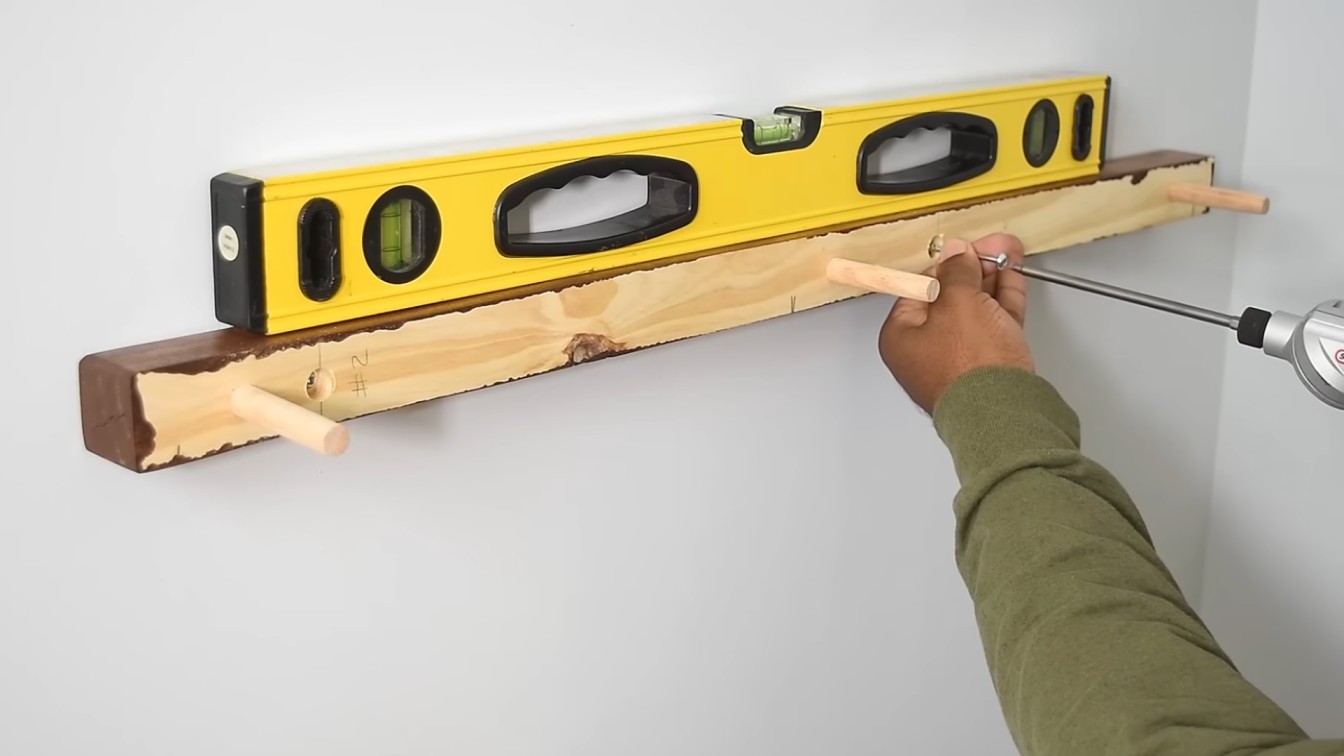 a person in a green sleeve uses a drill to hang a wooden shelf. A yellow level on top ensures it is straight