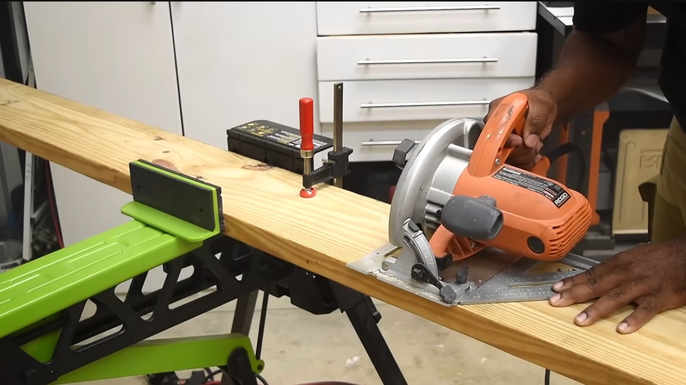 a person uses an orange circular saw to cut a wooden plank secured with a clamp in a workshop