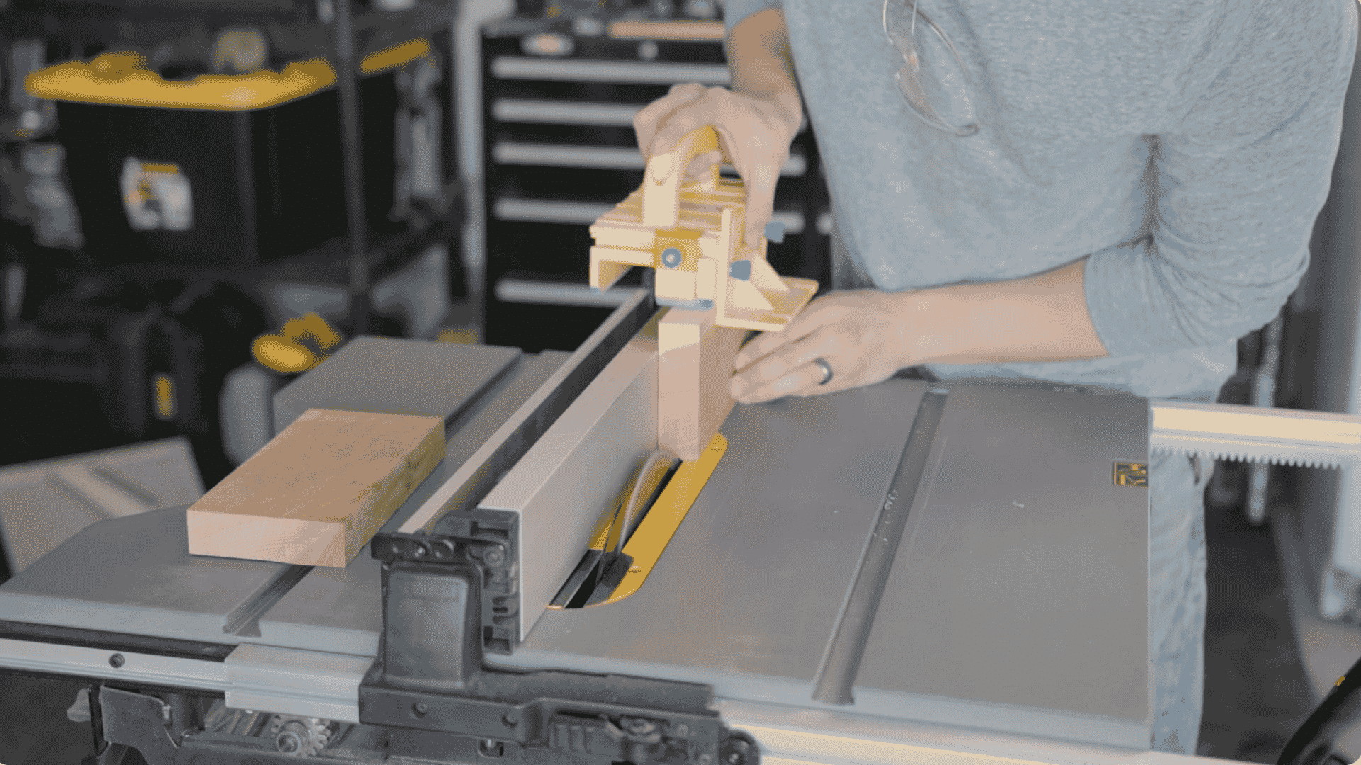 a person using a table saw with a push block to safely cut a wooden piece, with the saw blade exposed beneath the wood, the workspace includes various tools and storage in the background