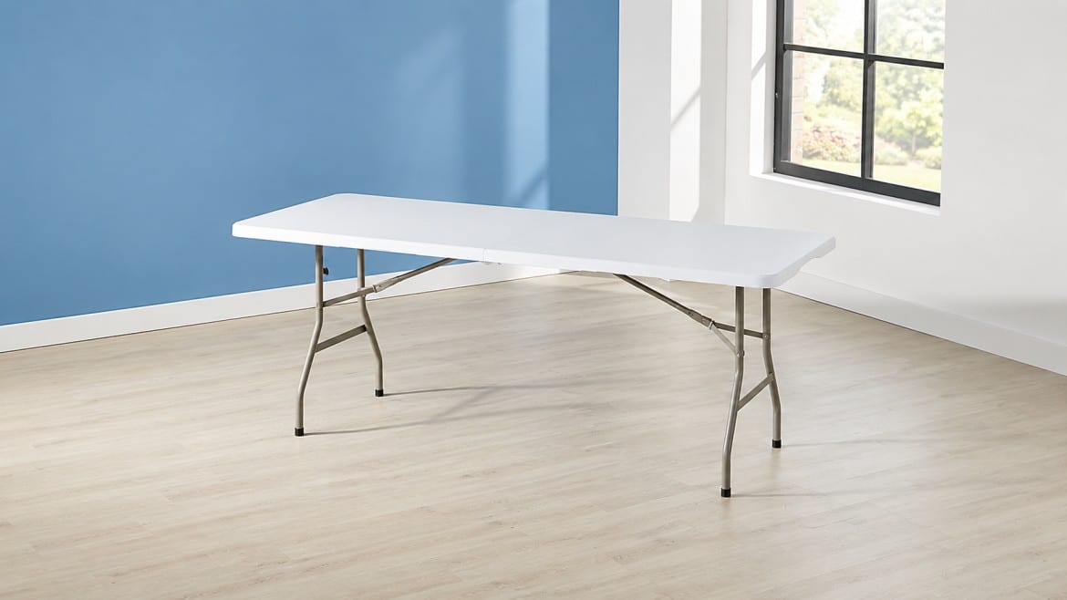 A white rectangular folding table with metal legs stands on a light wood floor, against a blue wall.