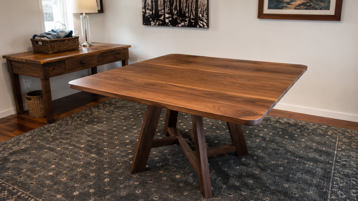 A wooden dining table with angled legs sits on a dark patterned rug