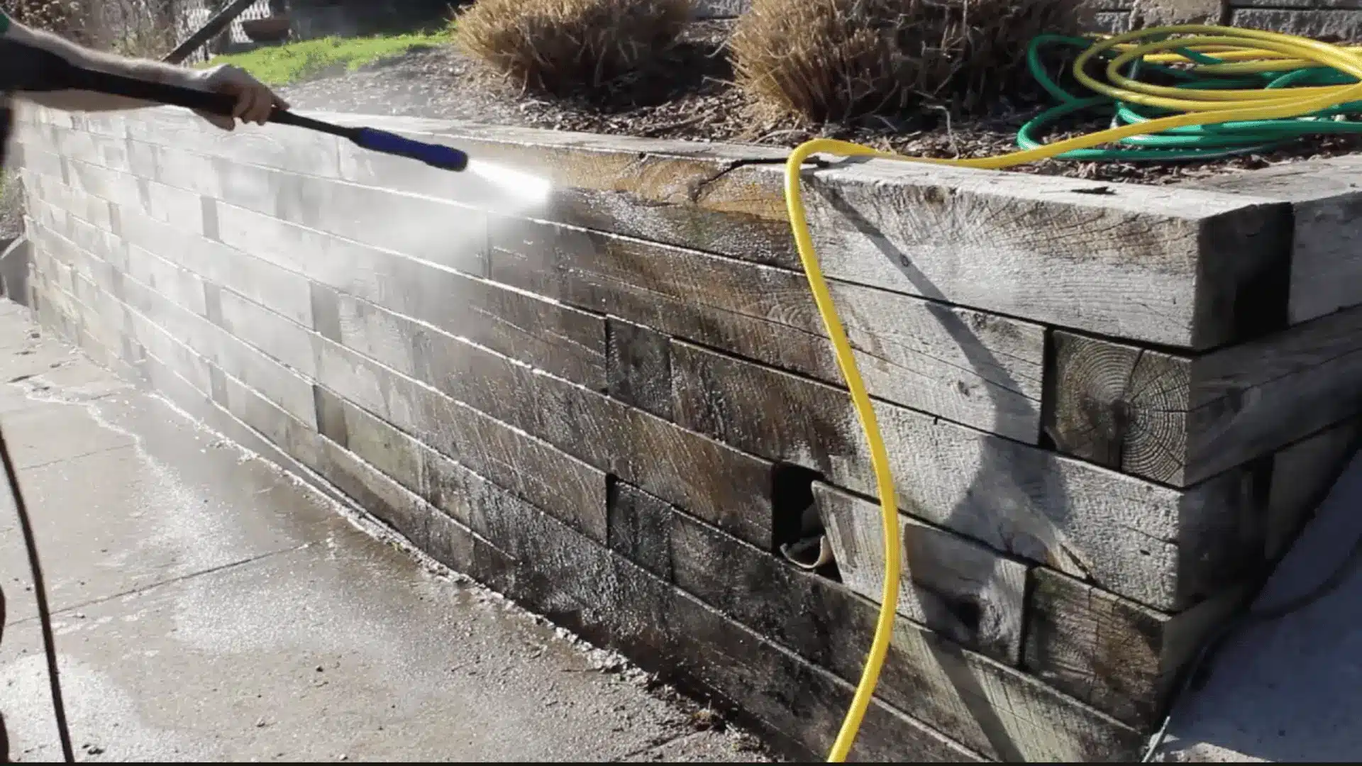 close-up photograph of pressure washing a wooden retaining wall made of stacked railroad ties with a yellow and green pipe