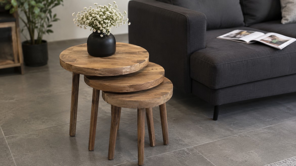 Nested wooden tables with a black vase of white flowers sit on gray tiles beside a dark gray sofa
