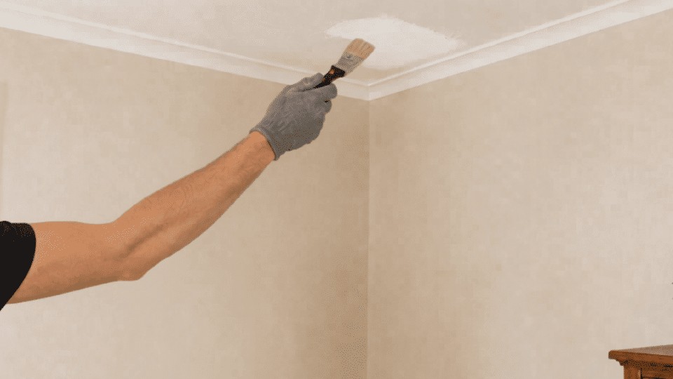 person applies stain blocking primer to the repaired ceiling corner with a brush beside a ladder