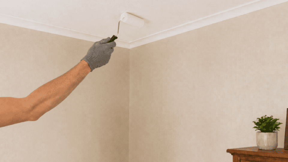 person repaints the repaired ceiling corner with a roller in the same room beside a ladder