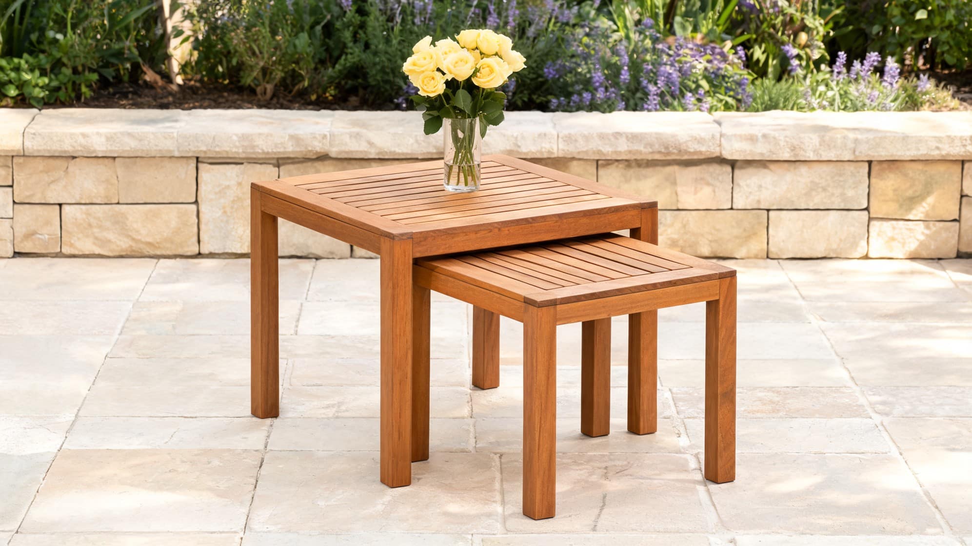 Two nesting wooden side tables on a patio with a vase of yellow roses