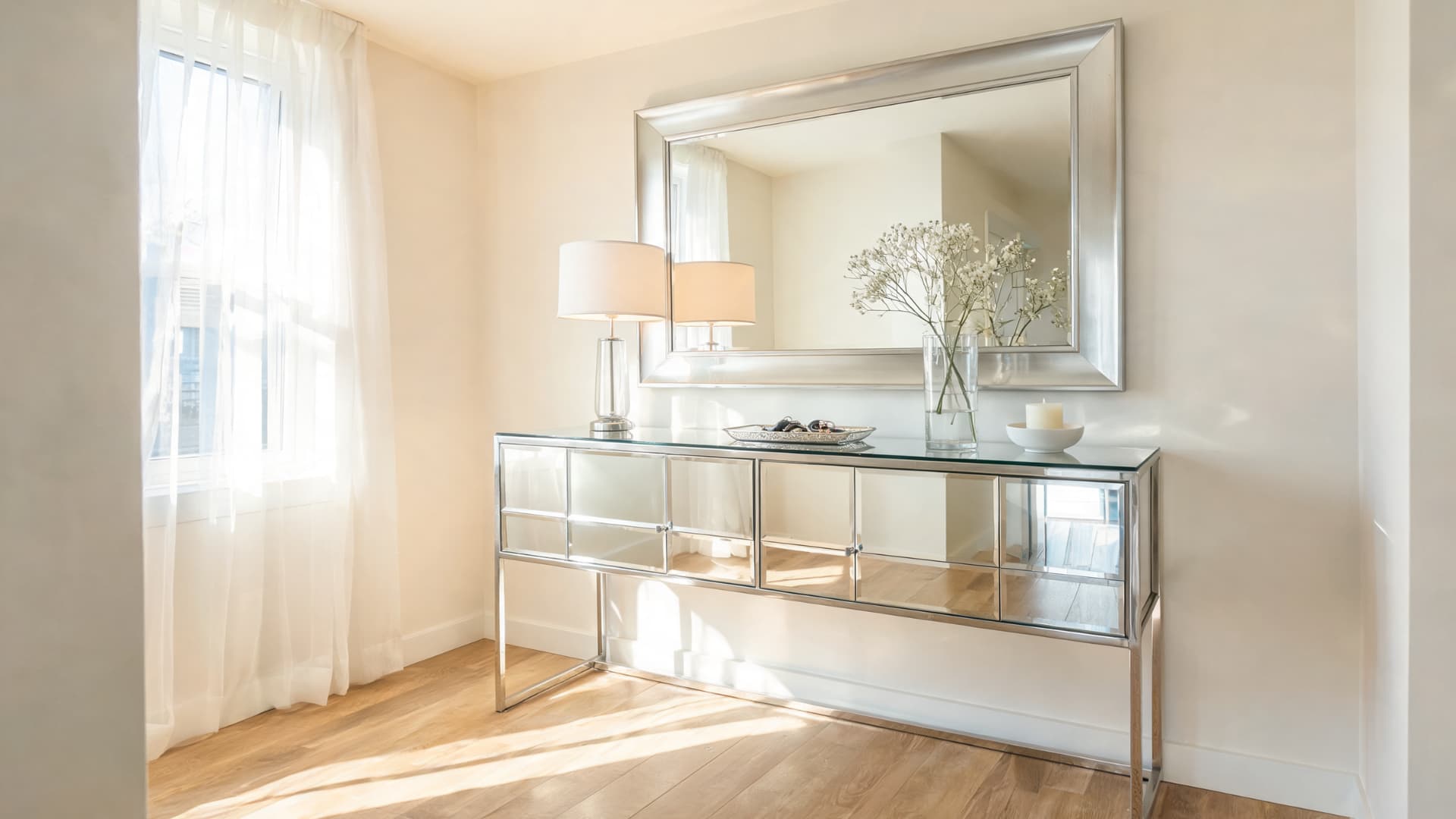 mirrored glass console table with reflective drawers, silver mirror, lamp, and bright sunlight in a small room