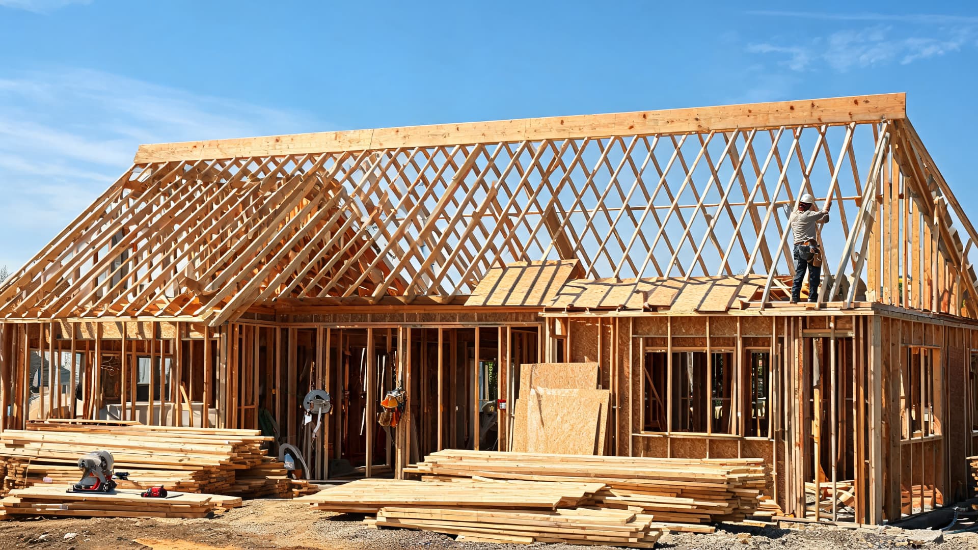 a new home frame with exposed roof rafters, lumber stacks, and a worker installing roof framing under a clear sky