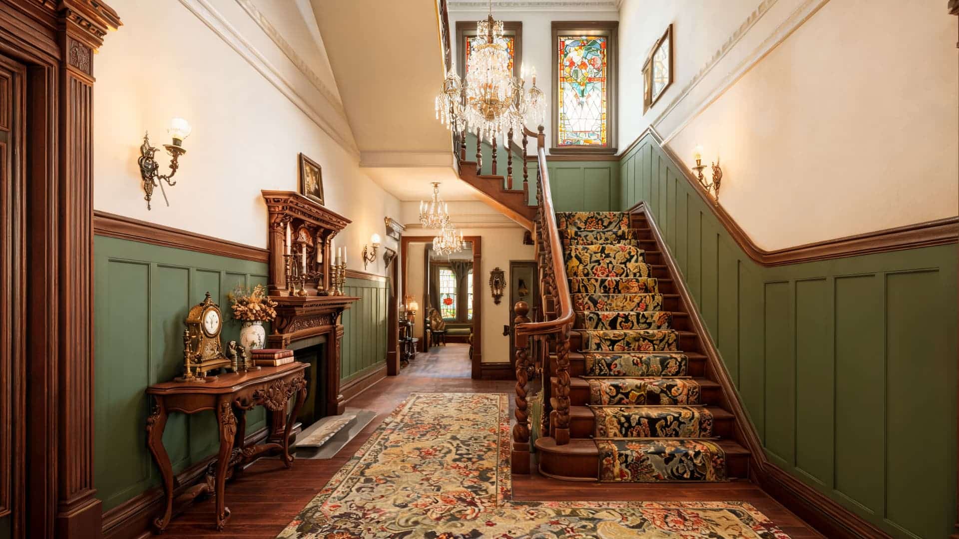 a victorian hallway with a carved wooden staircase green wainscoting and ornate rugs with chandeliers and stained glass windows