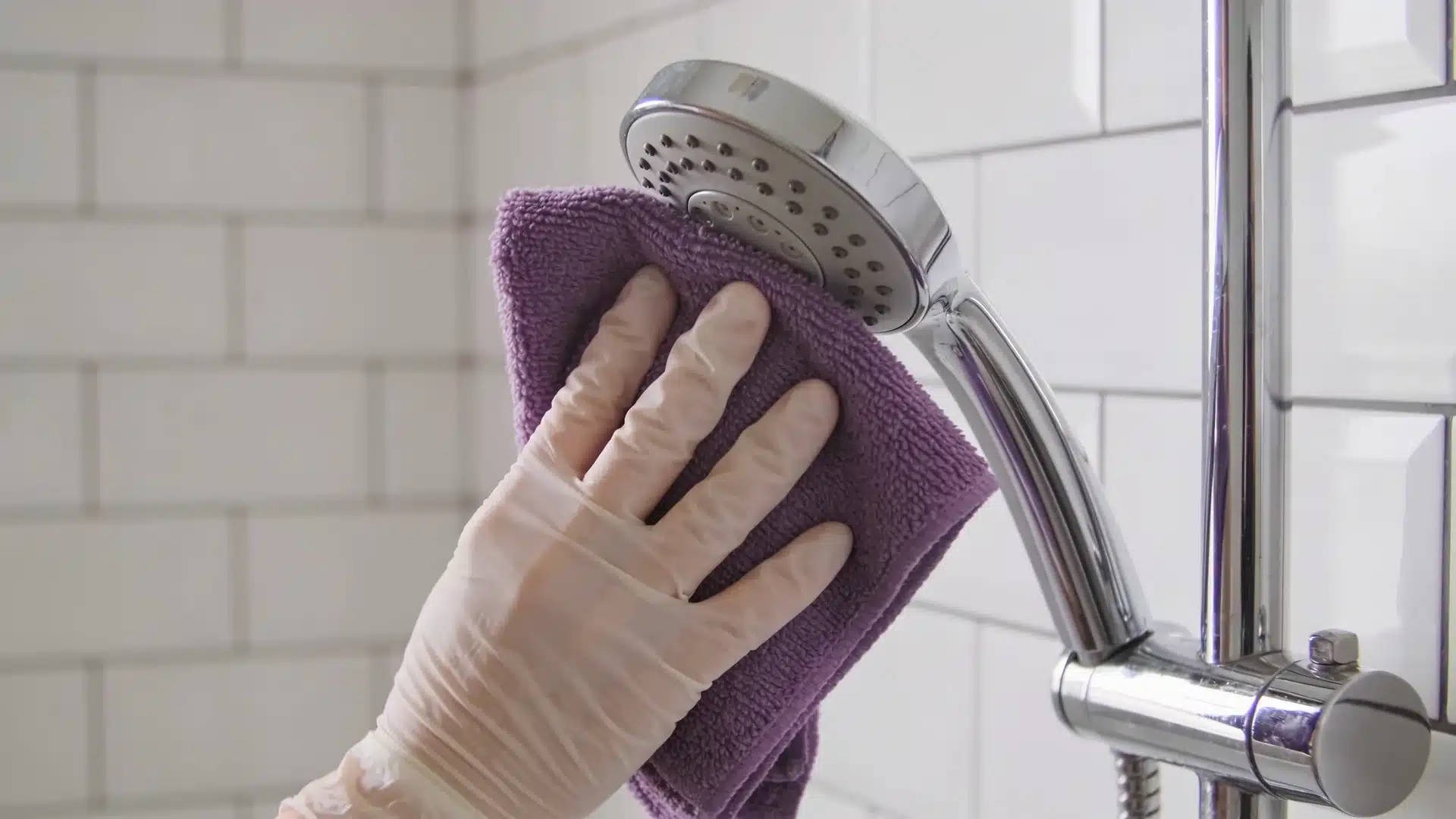 gloved hand wiping a chrome showerhead with a purple cloth against clean white subway tile