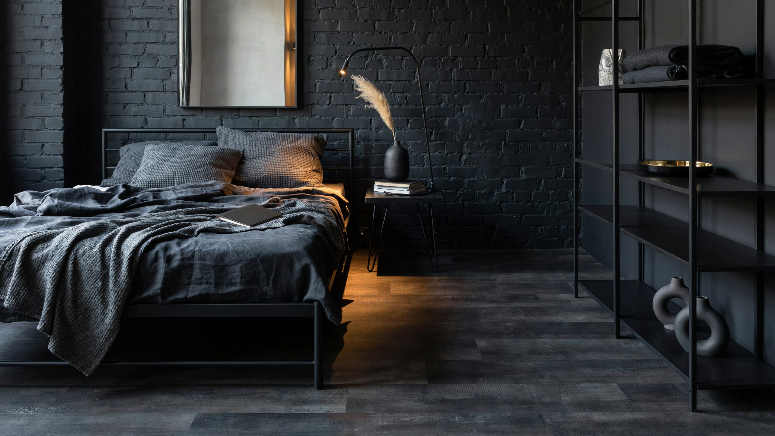 Dark bedroom with black metal bed frame, gray bedding, brick wall, shelving unit, and warm ambient lighting.