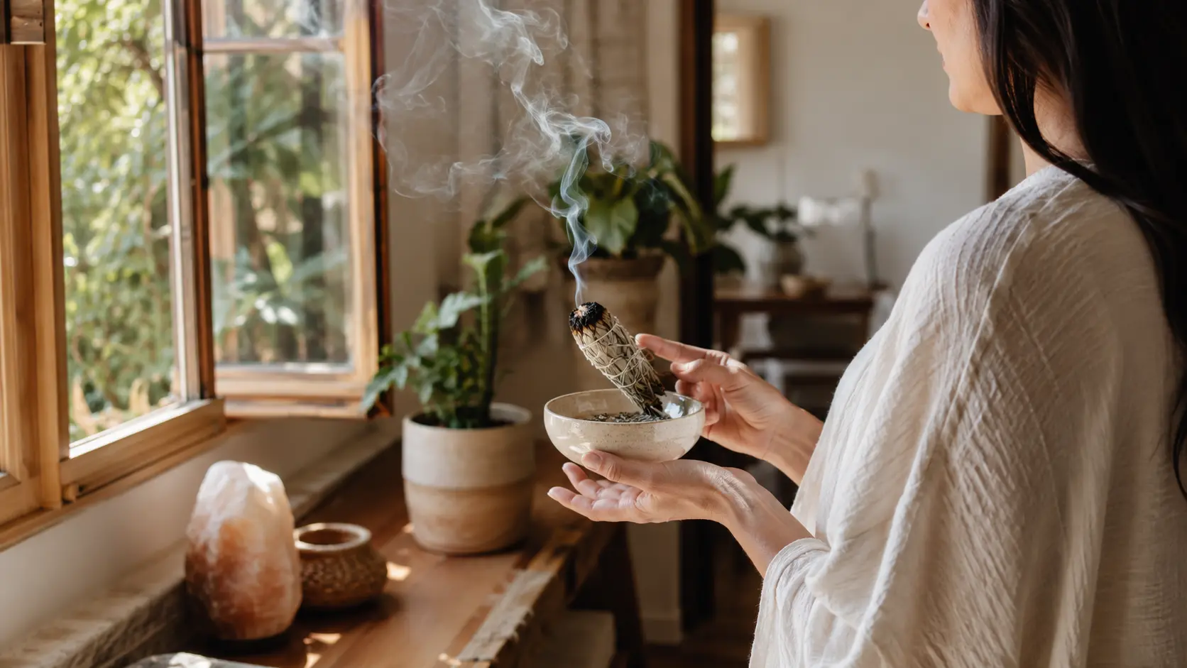 Person holding a smoking sage bundle over a ceramic bowl beside an open window in a warm, plant-filled room.