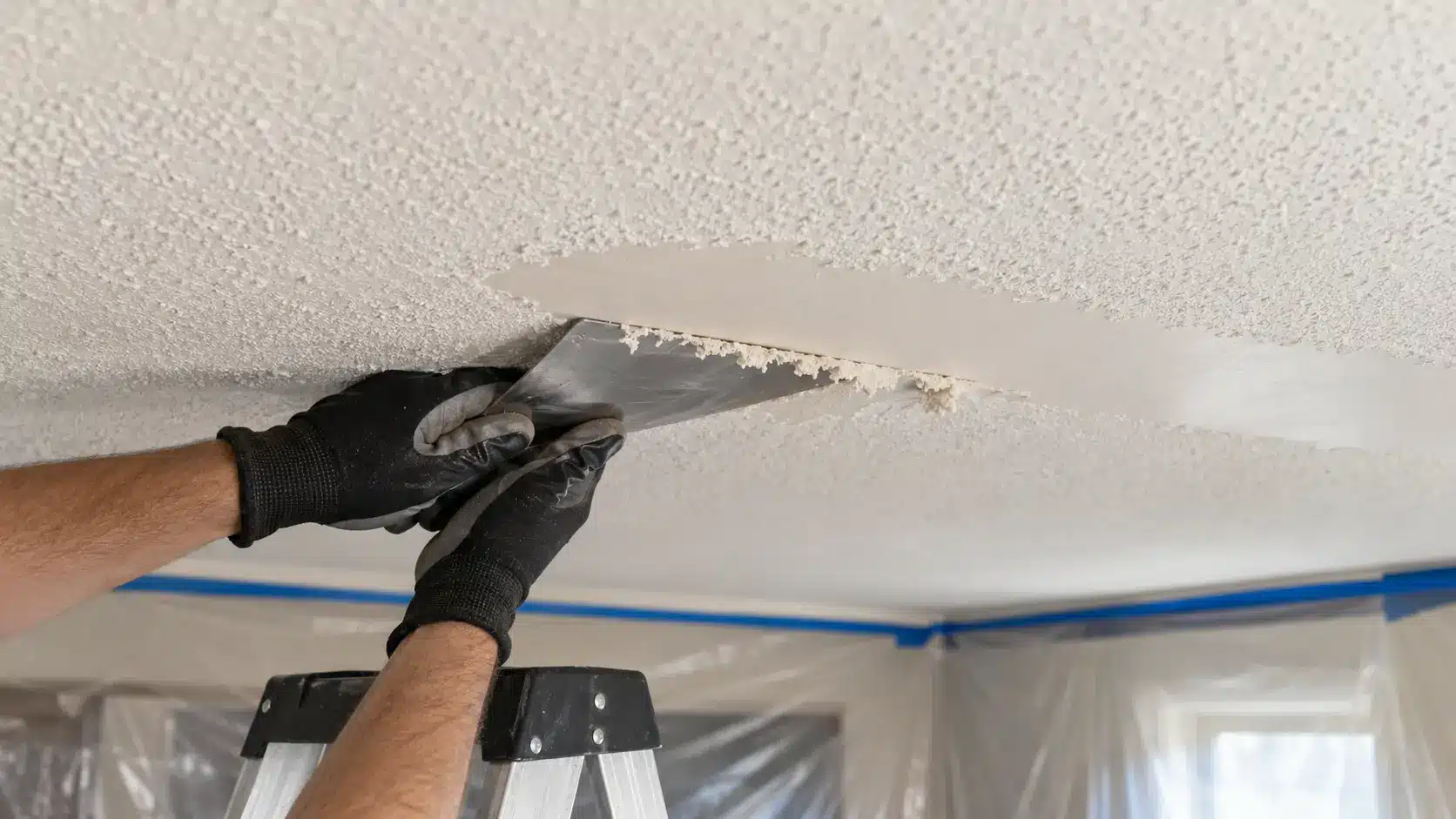 Gloved hands carefully scraping softened popcorn ceiling texture without damaging the drywall underneath.