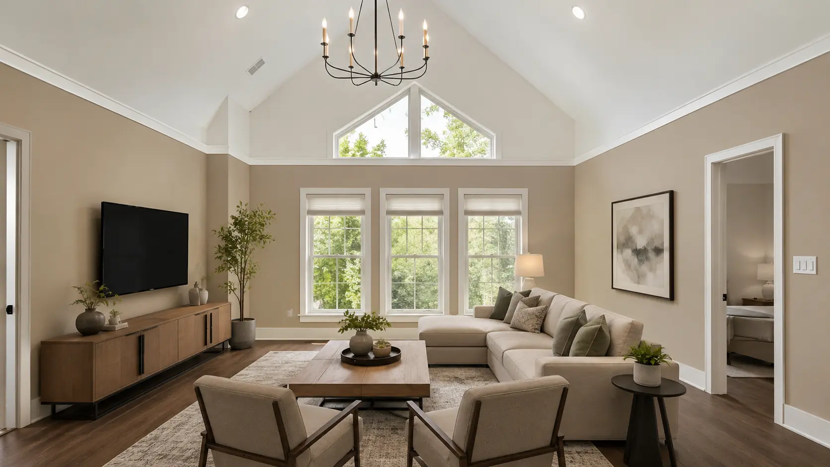 Modern living room with two-tone beige and white walls, a vaulted ceiling, black chandelier, large windows, neutral sectional sofa, and wall-mounted TV.